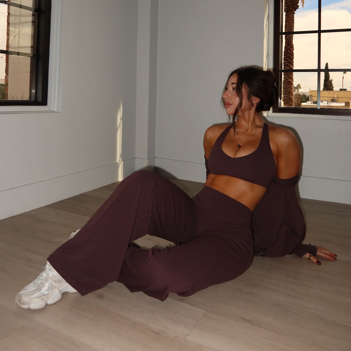 The coach sitting on a wooden floor in a brown matching set and white sneakers, looking toward a window.
